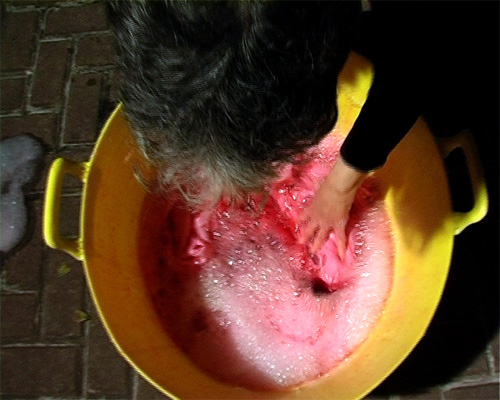 Video still: A woman washes a piece of material in a yelloe bucket filled with red liquid.