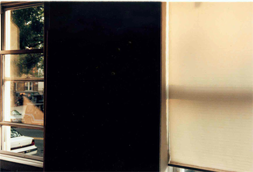 film still: Interior of a room with two wiindows looking out onto the street. One window had a blind pulled down.